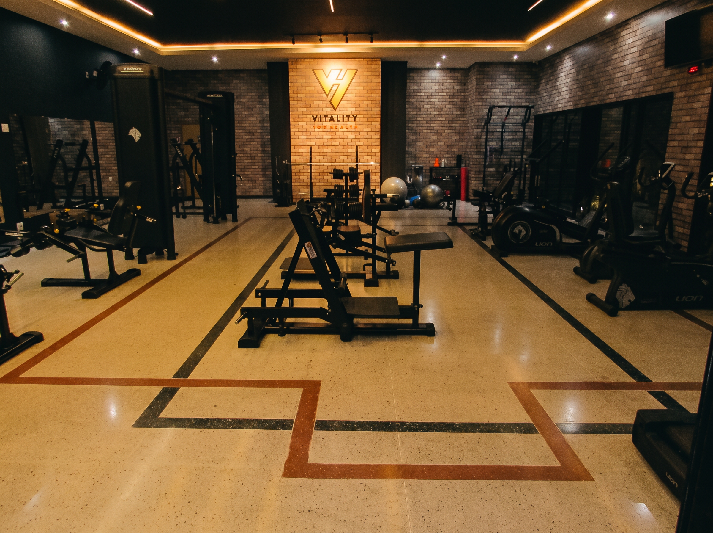 Salão principal do studio com leg press, bancos e a parede iluminada da logo Vitality for Health