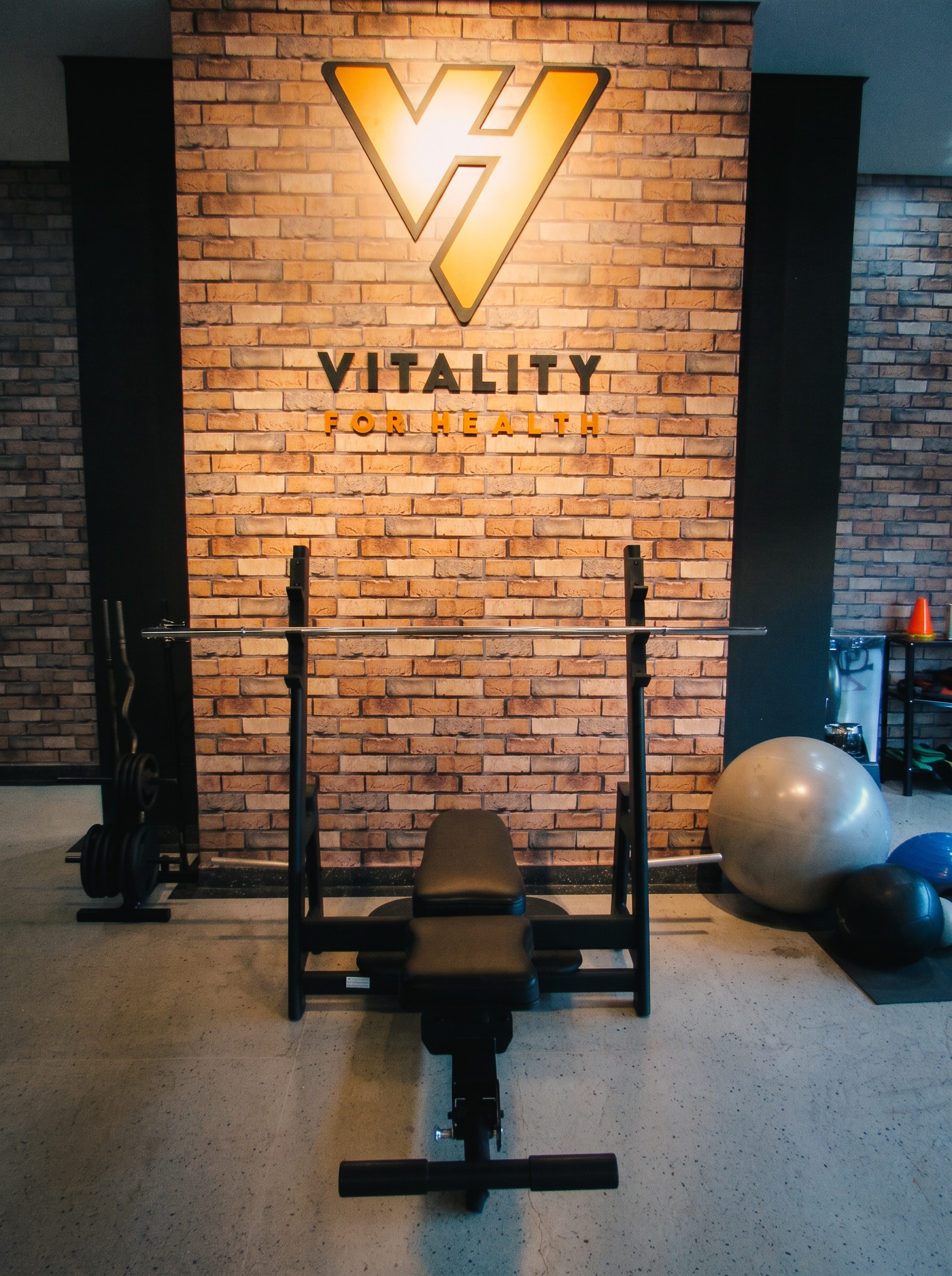 Banco de supino com barra e a parede da logo Vitality for Health iluminada ao fundo